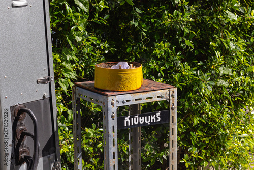 Foto Makeshift yellow outdoor ashtray on a metal stand with a Thai sign indicating a smoking area, placed next to an electrical utility box against lush green foliage