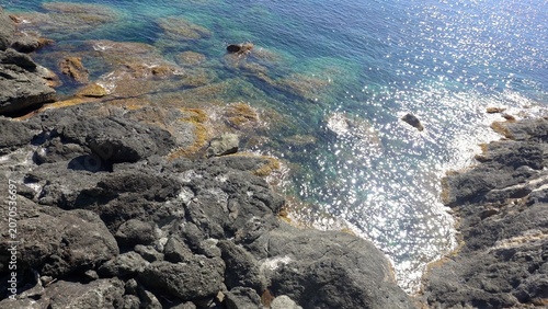 Clear, crystalline water on the Italian Mediterranean coast—Framura, Liguria, near the Cinque Terre and the Gulf of Tigullio—featuring a volcanic coastline, azure waters, and sun glints in summer