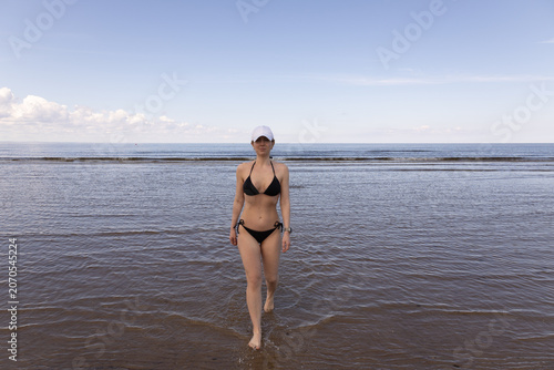 Young woman in a black bikini walking out of the calm sea during summer vacation. Healthy lifestyle, beach leisure and wellness concept on a sunny day.
