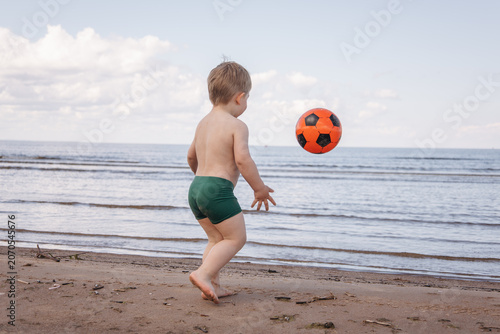 Toddler playing with a soccer ball on a sandy beach by the sea during summer vacation. Rear view of a child enjoying outdoor family leisure by the coast.