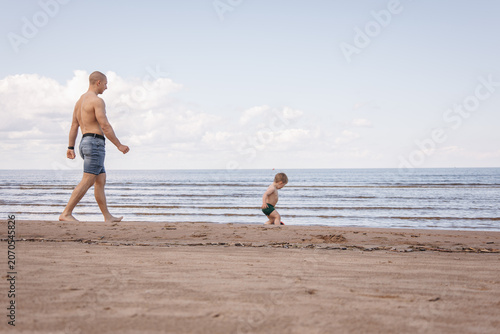 Father walking behind his little son on a sandy beach near the sea. Family spending quality time together outdoors during a peaceful summer day with copy space.