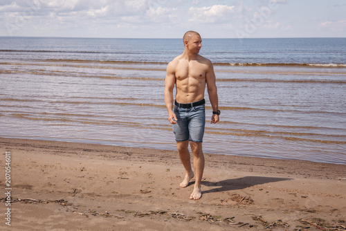 Shirtless man walking barefoot along a sandy beach by the sea on a sunny summer day. Rear view of an active adult enjoying a peaceful seaside walk during vacation.