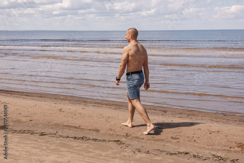 Shirtless man walking barefoot along a sandy beach by the sea on a sunny summer day. Rear view of an active adult enjoying a peaceful seaside walk during vacation.