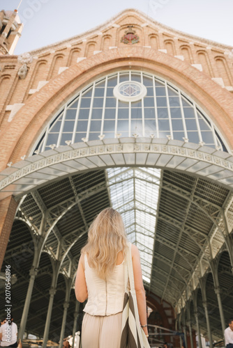 Young woman standing with her back under the historic Central Market building in Valencia, Spain. Female traveler exploring famous architecture and popular tourist destination during summer vacation.