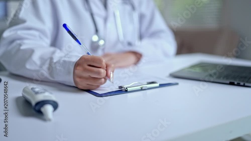 Close up of a female doctor in a white coat with a stethoscope writing a medical prescription or filling out a patient's health insurance form at her desk in a modern hospital clinic