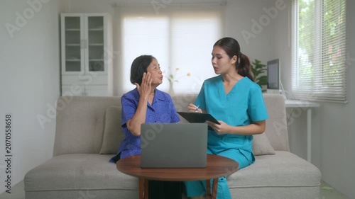 Young Asian female caregiver wearing scrubs talking with an elderly woman, explaining health information and taking notes on a clipboard during a home visit in a living room environment