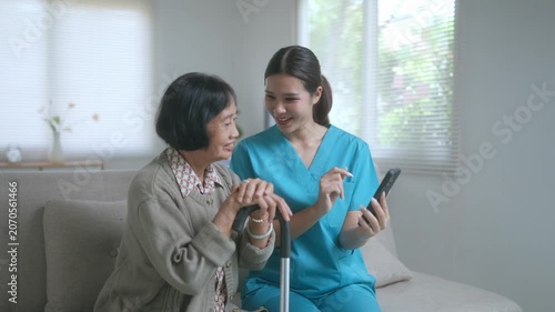 Professional caregiver teaching a senior patient how to use a mobile phone application while sitting together on a comfortable sofa in a bright, modern living room at a nursing home