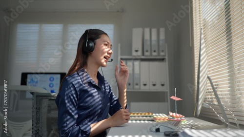 Young Asian business woman wearing a headset communicating with clients via video conference, providing online customer support and telemarketing services in her modern office workplace