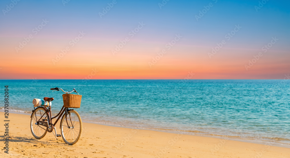 Fototapeta premium Vintage bicycle with a wicker basket parked on a sandy beach by a turquoise sea at sunset