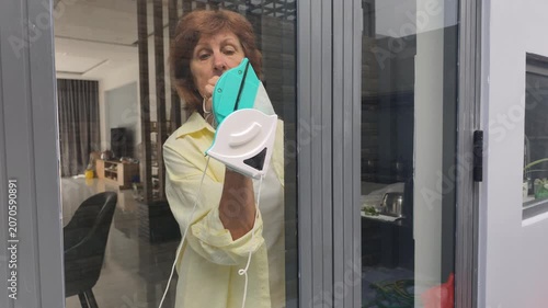 Tired woman struggles with heavy magnetic device during window cleaning process. Hard manual labor and awkward movements make window cleaning difficult, leaving streaks on dirty apartment glass.