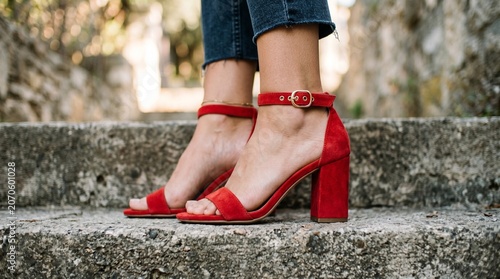 A woman wears vibrant red suede block heel sandals with gold buckles and denim jeans while standing gracefully on textured stone steps in soft natural lighting.