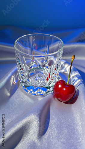 Crystal glass and red cherry on blue satin fabric.