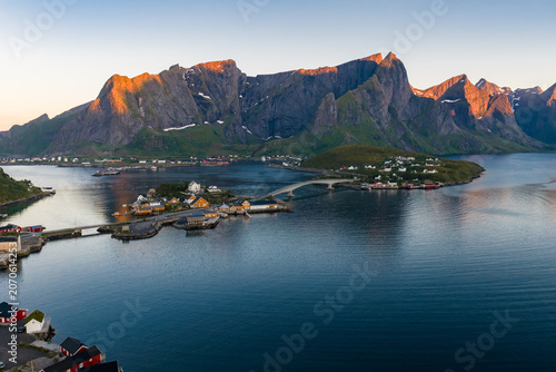 Tableau sur toile Aerial summer scene of Reine, Lofoten, Norway, with Reinebringen peaks lit by evening sun, rorbuer cabins along the shore, arched bridges linking islets over a calm fjord