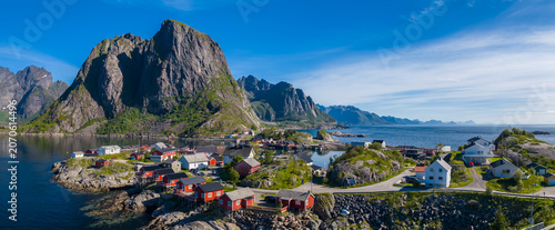 Foto Aerial view of a Lofoten Islands village in Norway with red rorbuer, white houses, rocky islets, calm water, bridges, curving roads, and towering peaks in bright summer light