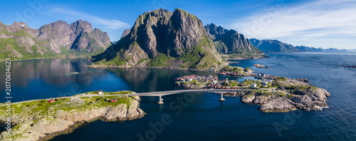 Aerial scene shows Reine and Reinebringen in Norway, granite peaks, deep fjords, a sleek bridge, red rorbuer cabins, calm turquoise shallows, and clear summer daylight.