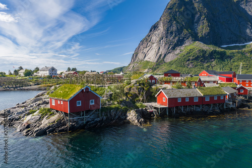 Принти на полотні Traditional rorbuer with grass roofs sit on rocky islets above clear turquoise water, beneath a granite peak in Lofoten, Norway, under midday summer sun