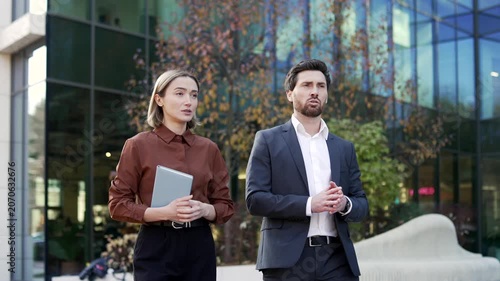Close-up of professional colleagues talking while walking near a modern office building. Businessman and businesswoman discuss ideas and business matters outdoors in a contemporary urban environment.
