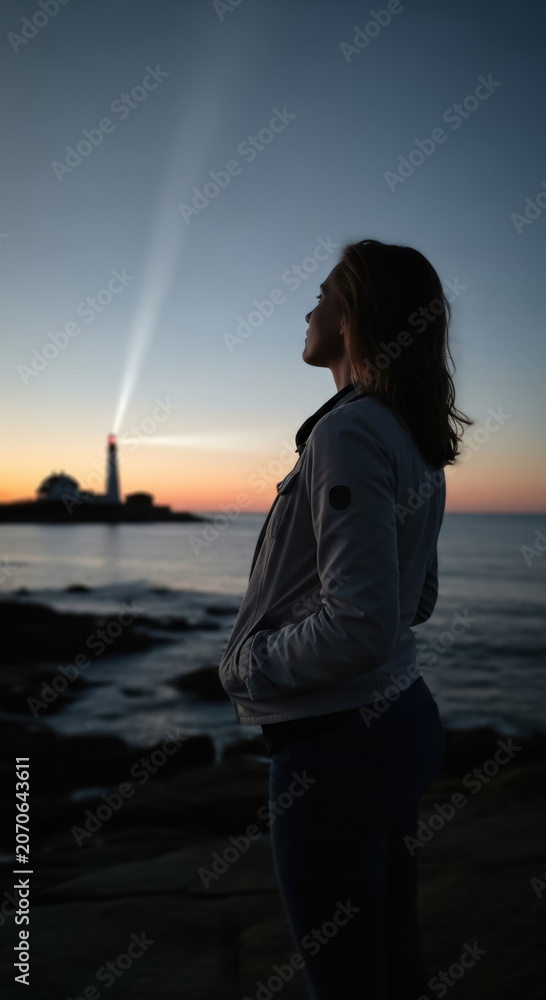 Fototapeta premium Woman watches a lighthouse beam cutting through the dusk sky while standing on a coastal rock formation.