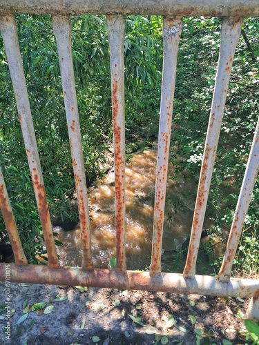 High angle view of a rushing brown muddy stream or river flowing rapidly below old rusty metal bridge bars after heavy rain.