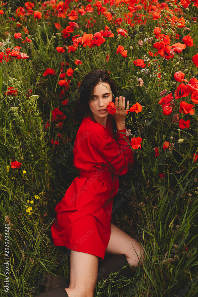 Naklejka premium Sensual brunette woman with a gentle sideways glance and open mouth posing genuinely in a vibrant poppy field, wearing an authentic local red wrap dress with boots, cinematic lifestyle portrait
