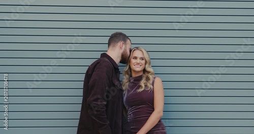 An emotional shot of a loving couple standing against a modern gray wall. The man gently kisses his woman, and she smiles sincerely.