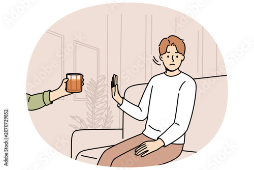 Determined man refuses alcohol, demonstrates stop gesture, sitting on sofa near hand with whiskey. Guy says no to alcohol and wants to live sober lifestyle to avoid hangover and intoxication