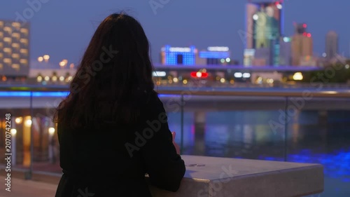 Woman with long dark hair sits alone overlooking illuminated cityscape, river, buildings reflected in water during twilight hour with blue, golden lights creating serene urban atmosphere.