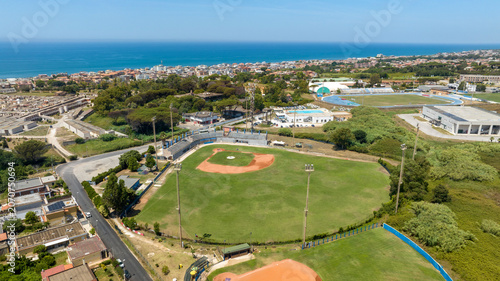 Φωτογραφία Aerial panorama of a beautiful baseball field nestled within a coastal sports complex, showcasing the green outfield, red dirt infield, and a stunning blue sea horizon