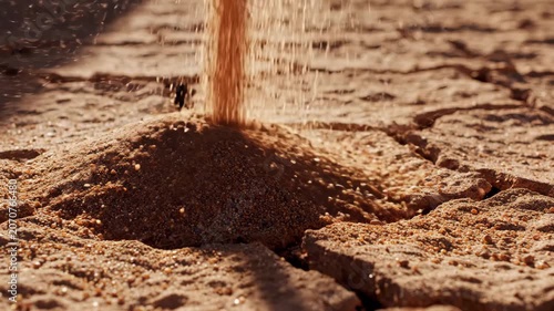 Reddish-brown sand pouring onto dry cracked earth surface forming a small mound.