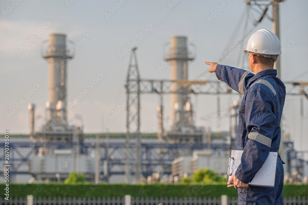 Fototapeta premium Asian male electrical engineer holding digital tablet standing at power plant station. Professional energy industry technician working on infrastructure maintenance and renewable resource project.