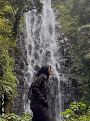 Muslim Woman in Black Hoodie and Hijab Standing Near Waterfall in Forest Landscape. Muslim woman wearing a black hoodie and black hijab while standing near a waterfall in a tropical forest landscape, 