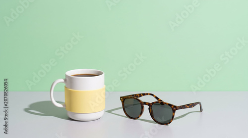 Ceramic coffee mug and tortoiseshell sunglasses in contemporary aesthetic layout in pastel green and neutral grey palette representing summer morning routine with copy space