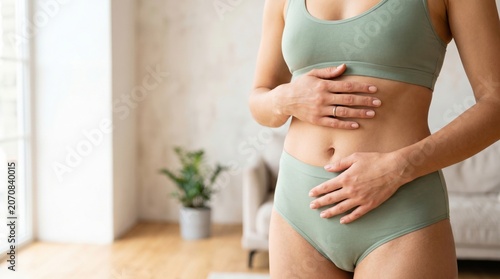 A woman stands in a bright room with a plant nearby. She has her hands on her stomach while wearing simple clothing. Sunlight fills the space, creating an open feel