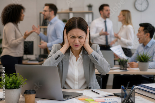 A woman sits at her desk with her hands over her ears. She looks stressed while other workers talk loudly and argue around her. Papers and a laptop are on her desk.