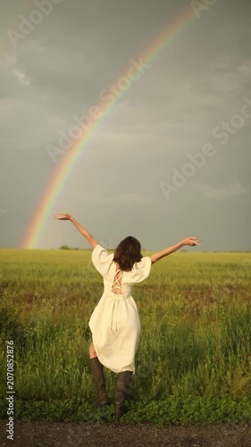 Brunette woman from back view standing in green field with arms raised to spectacular vibrant rainbow, joyful celebration rainbow miracle majestic summer nature stormy sky lifestyle video footage