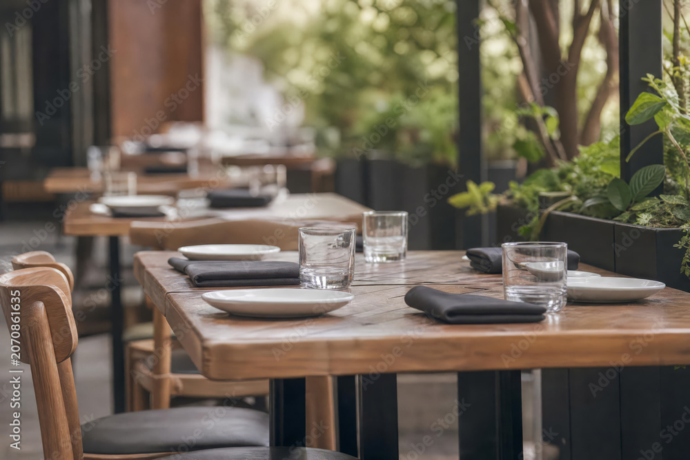 Fototapeta premium Side Angle Restaurant Patio Table (No Food) With Glassware And Greenery Photographed In Bright Commercial With Layered Restaurant Depth