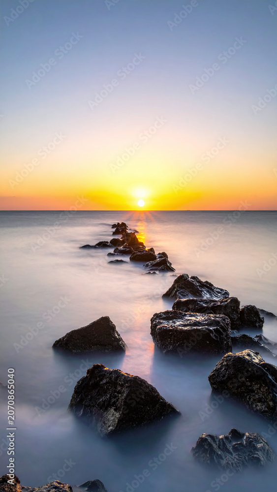 Fototapeta premium This tranquil coastal scene features a rocky path stretching into a golden sunset over silky water. A peaceful metaphor for a journey, perfect for wellness, meditation, and travel designs.