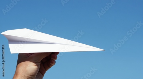 Hand launches white paper airplane against clear blue sky. Simple origami toy flies outdoor. Childhood fun activity, creative gesture. Travel, dreams, freedom.