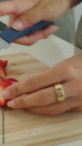 Vertical video: Gripping blue knife, cook in apron slicing strawberry at counter, preparing dish