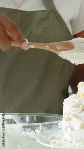 Vertical video: Lifting spoon, baker in olive apron scraping and decorating icing at counter, cake