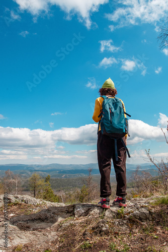 Young woman with backpack standing on cliff edge. Girl hiking on mountain top overlooking forest landscape under blue sky. Active lifestyle, adventure, travel and exploration concept.