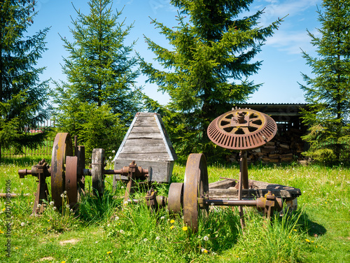 Canvas Print Rusty vintage farming equipment and iron wheels sit in a grassy meadow surrounded by evergreen trees under a bright blue sky