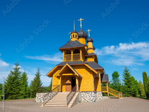 Obraz na plátne A beautiful yellow wooden Orthodox church with dark onion domes and a prominent bell tower stands on a paved square surrounded by green trees on a bright sunny day
