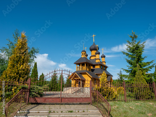 Quadro em tela An ornate brown metal gate stands at the entrance of a courtyard leading to a traditional yellow wooden Orthodox church with onion domes