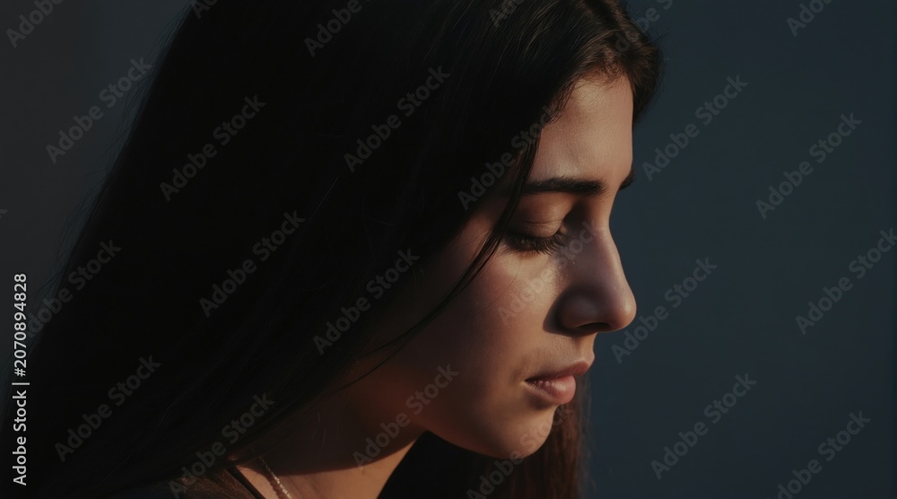 Fototapeta premium Young woman looks down with closed eyes in shadow. Soft light on face suggests sadness or deep thought. Her gaze is introspective, conveying quiet melancholy.