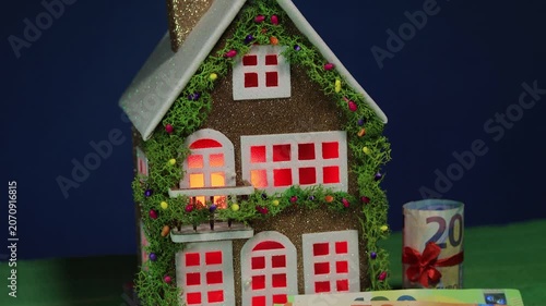 On a dark blue background and green covering, a decorated house with color-changing light in the windows and euro banknotes nearby