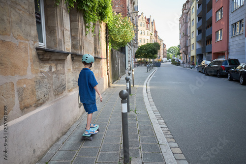 Young boy in blue outfit rides a skateboard down a city sidewalk