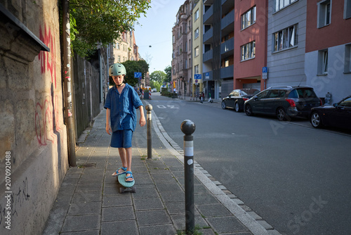 Young boy in blue outfit and helmet rides a skateboard down a city sidewalk