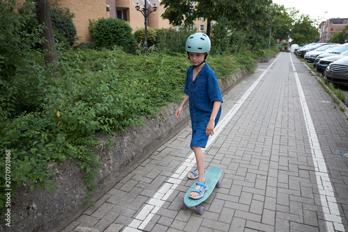 Young boy in blue outfit and helmet rides a skateboard on a paved path
