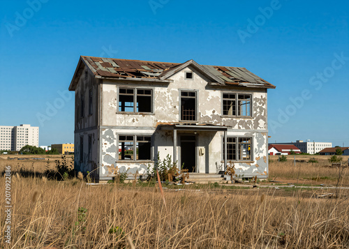 An abandoned house with peeling paint and broken windows stands alone in the middle of dry grass, with modern buildings visible in the distance